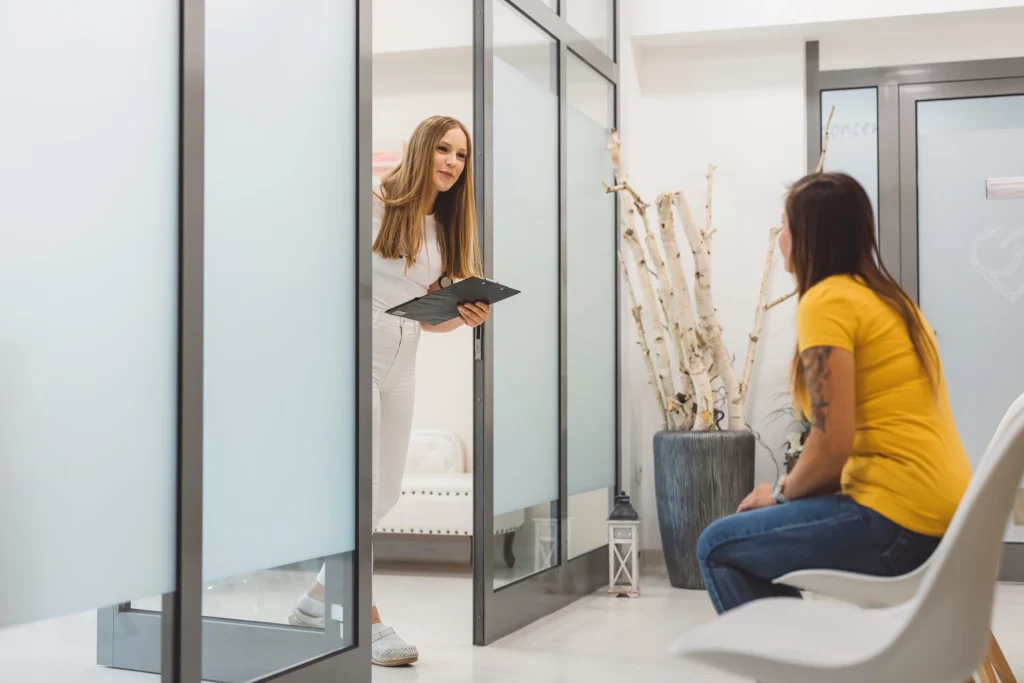 Staff member with a clipboard opens a frosted glass door beside a seated visitor in a waiting area.